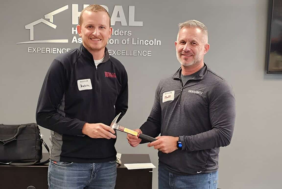 Two men smiling, one passing a hammer, HBAL logo background.