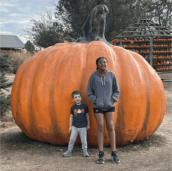 Two children stand in front of a giant pumpkin at a fall festival.