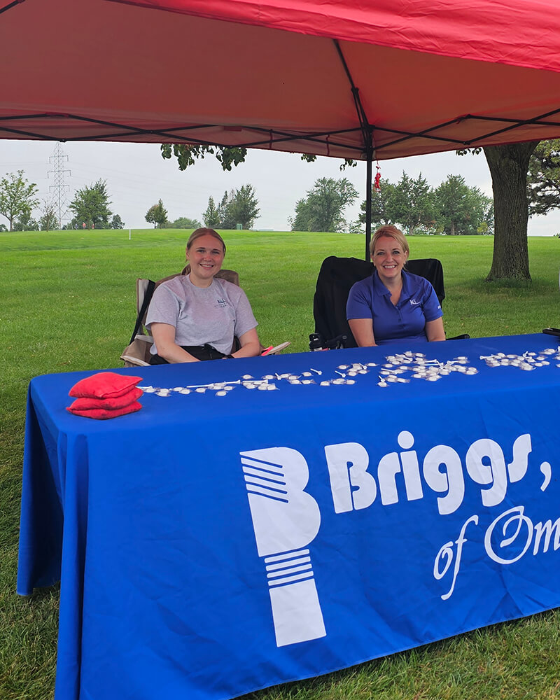 Two smiling women behind a Briggs table at outdoor event.