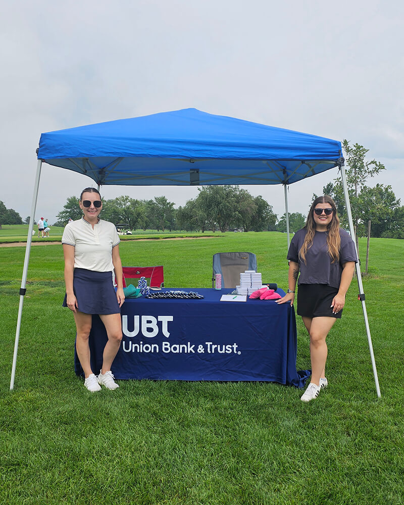 Two women smiling at UBT booth on golf course.