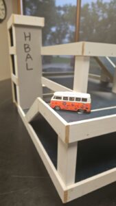 Small toy bus on a white wooden model structure with 'H BAL' written on it.
